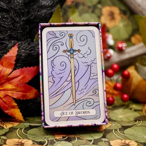 May include: A tarot card from the Ace of Swords deck, featuring a golden sword against a backdrop of purple and white clouds. The card is in a decorative box with a floral pattern.