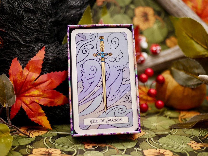 May include: A tarot card from the Ace of Swords deck, featuring a golden sword against a backdrop of purple and white clouds. The card is in a decorative box with a floral pattern.