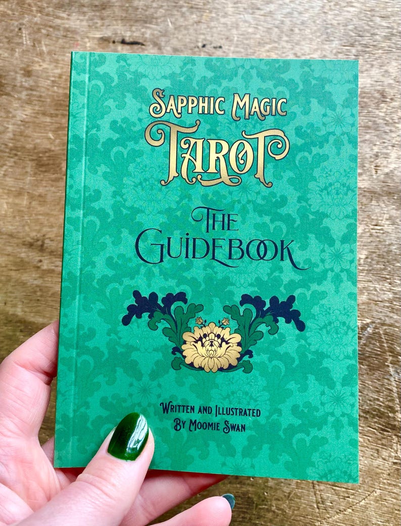 May include: A green tarot guidebook titled "Sapphic Magic Tarot: The Guidebook." The cover features ornate gold lettering and a floral design. The text "Written and Illustrated by Moomie Swan" is also visible.