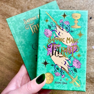 May include: Two tarot card decks with teal covers. The front deck features the words "Sapphic Magic Tarot" in gold script, with illustrations of hands, flowers, and a chalice. The other deck is visible in the background. The decks are by "Moomie Swan."