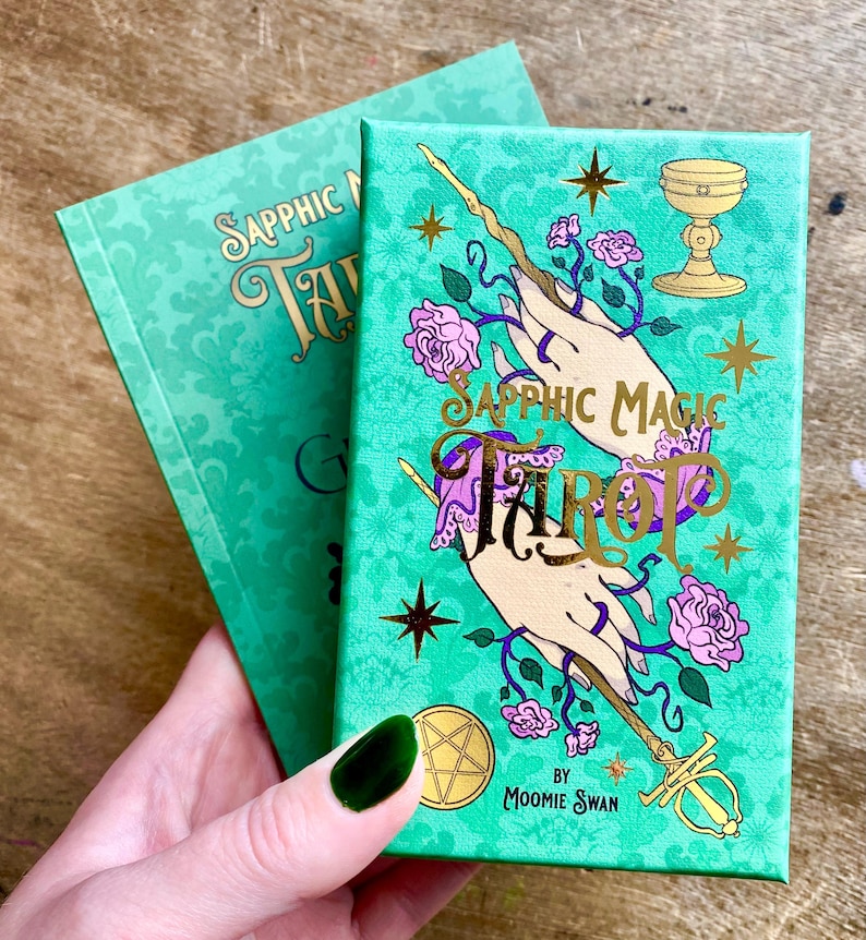 May include: Two tarot card decks with teal covers. The front deck features the words "Sapphic Magic Tarot" in gold script, with illustrations of hands, flowers, and a chalice. The other deck is visible in the background. The decks are by "Moomie Swan."