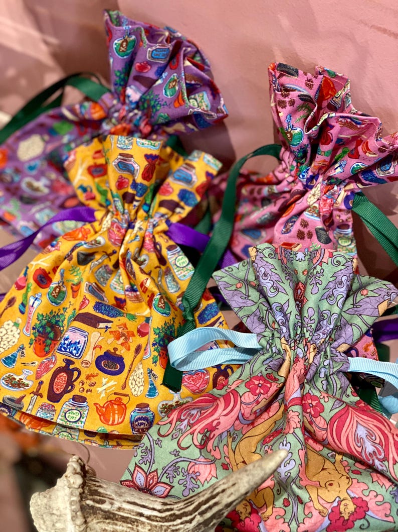 May include: A collection of colorful drawstring bags with various patterns. The bags feature designs with jars, teapots, and floral motifs. Each bag is tied with a contrasting ribbon, including green, purple, and blue.