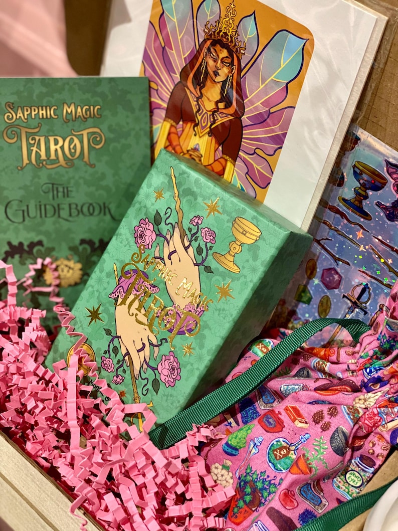 May include: A gift box containing a green tarot card box with gold lettering and illustrations, a green guidebook, and a colorful illustration of a woman with wings. The box is filled with pink paper filler and a pink patterned fabric.