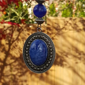 May include: A blue glass pendant with a silver metal frame and a silver chain. The pendant is oval shaped and has a swirl design. The chain has a silver clasp.