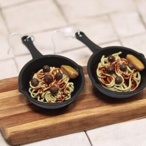 May include: Two miniature black cast iron skillets with handles, each containing a small portion of spaghetti with meatballs and a slice of bread. The skillets are on a wooden cutting board.