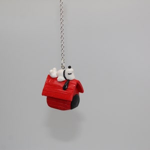 May include: A Snoopy-themed ceiling fan pull featuring a red doghouse with a sleeping Snoopy on top. The doghouse has a black door and a silver chain for hanging. The Snoopy figure is white with black accents.