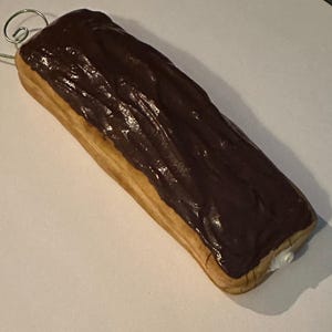 May include: A long, rectangular pastry with a light brown base and a thick, glossy dark chocolate topping. A small amount of white cream is visible at one end. A silver wire loop is attached to the top.