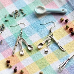 May include: A pair of silver-toned dangle earrings featuring miniature cutlery charms, including a knife, fork, and spoon on each hook. The earrings are displayed alongside a small light blue ceramic measuring spoon, a metal whisk charm, and scattered dried lavender buds and peppercorns.