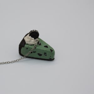 May include: A green mint chocolate chip pie slice with a chocolate cookie on top, hanging from a silver chain. The pie slice is made of polymer clay.