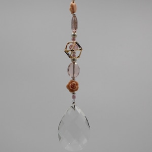 May include: A pink and gold crystal suncatcher with a teardrop-shaped crystal, a geometric bead, and pink flower accents.
