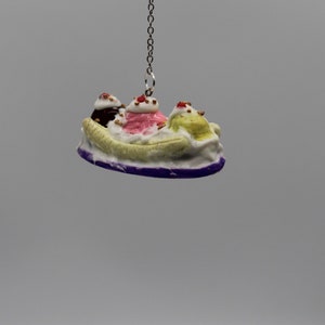 May include: A banana split charm with three scoops of ice cream, chocolate, strawberry, and pistachio, topped with whipped cream and cherries, hanging from a silver chain.