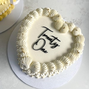 May include: A heart-shaped cake with white icing and a piped border. The cake is decorated with the words "Twenty One" in black script and gold sprinkles. Another cake with yellow icing and white ribbon is in the background.