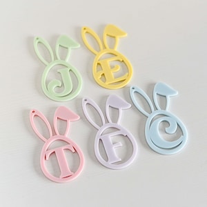 May include: Six pastel-coloured Easter bunny ornaments with letters cut out. The letters spell out the word "JEFT" in a light green, yellow, lavender, pink, and light blue colour scheme.