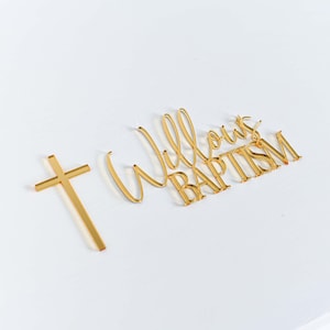 May include: Gold-colored baptism decorations on a white surface. The decorations include a cross and the words "Willow's Baptism" in a cursive font. The image is well-lit, highlighting the metallic sheen of the decorations.