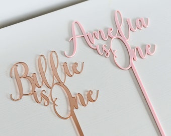 Puede incluir: Dos adornos para tartas, uno rosa y otro rosa dorado, con los nombres Amelia y Billie y el texto "is one" en cada uno.