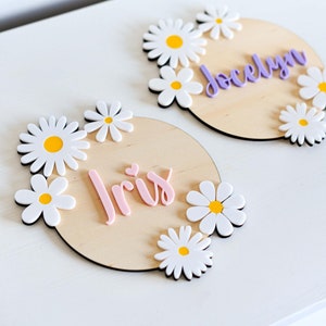 Può includere: Due cartelli di legno con fiori di margherita bianchi e nomi scritti in lettere rosa e viola. I nomi sono "Iris" e "Jocelyn".