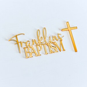 May include: Gold-colored baptism cake topper with the name "Franklin's" and the word "BAPTISM" in a decorative font. A gold cross is positioned to the right. The items are on a white surface.