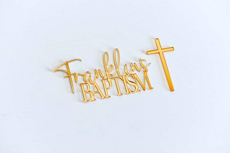 May include: Gold-colored baptism cake topper with the name "Franklin's" and the word "BAPTISM" in a decorative font. A gold cross is positioned to the right. The items are on a white surface.