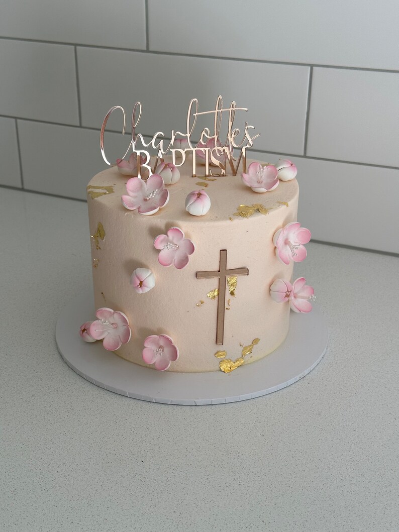May include: A round, light-colored cake decorated with pink flowers and gold leaf accents. The cake has a cross and the words "Charlotte's BAPTISM" in gold lettering. The cake sits on a white cake board.
