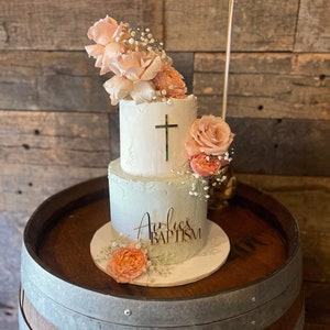 May include: A two-tiered white cake decorated with peach-colored roses and baby's breath. A gold cross is on the top tier, and the bottom tier has the words "Aurlie's BAPTISM" in gold script. The cake sits on a white cake board.