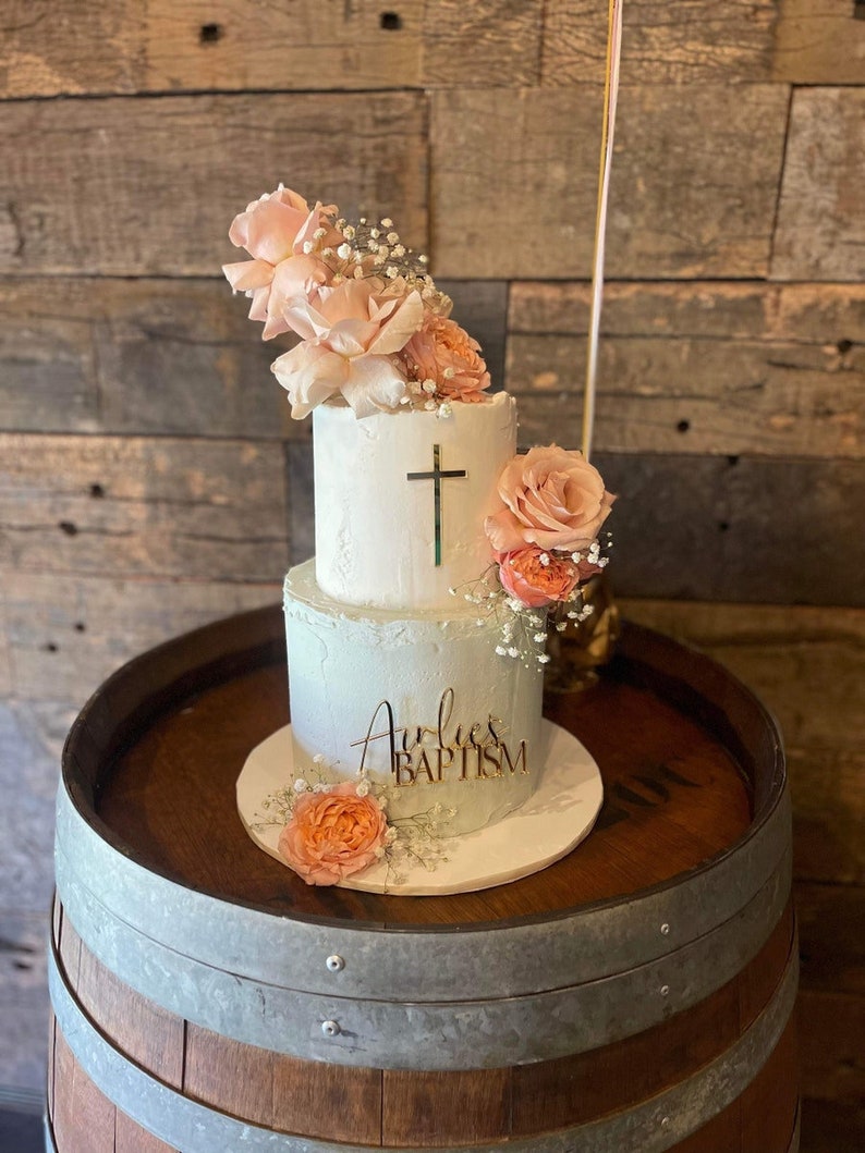 May include: A two-tiered white cake decorated with peach-colored roses and baby's breath. A gold cross is on the top tier, and the bottom tier has the words "Aurlie's BAPTISM" in gold script. The cake sits on a white cake board.