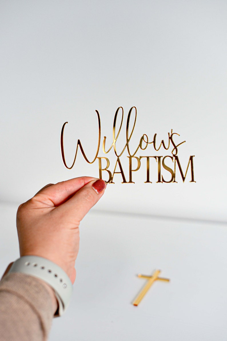 May include: A gold-colored, cursive-style sign that reads "Willow's BAPTISM" is held up by a hand with a light-colored watch. A small gold cross is in the background. The sign is likely for a baptism celebration.
