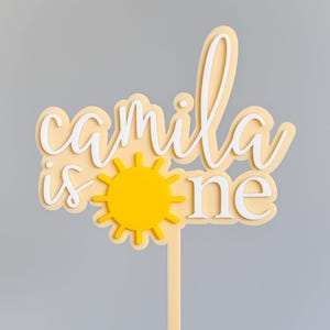 May include: A yellow and white cake topper with the text "Camila is one" and a yellow sun graphic. The topper is on a beige stick.