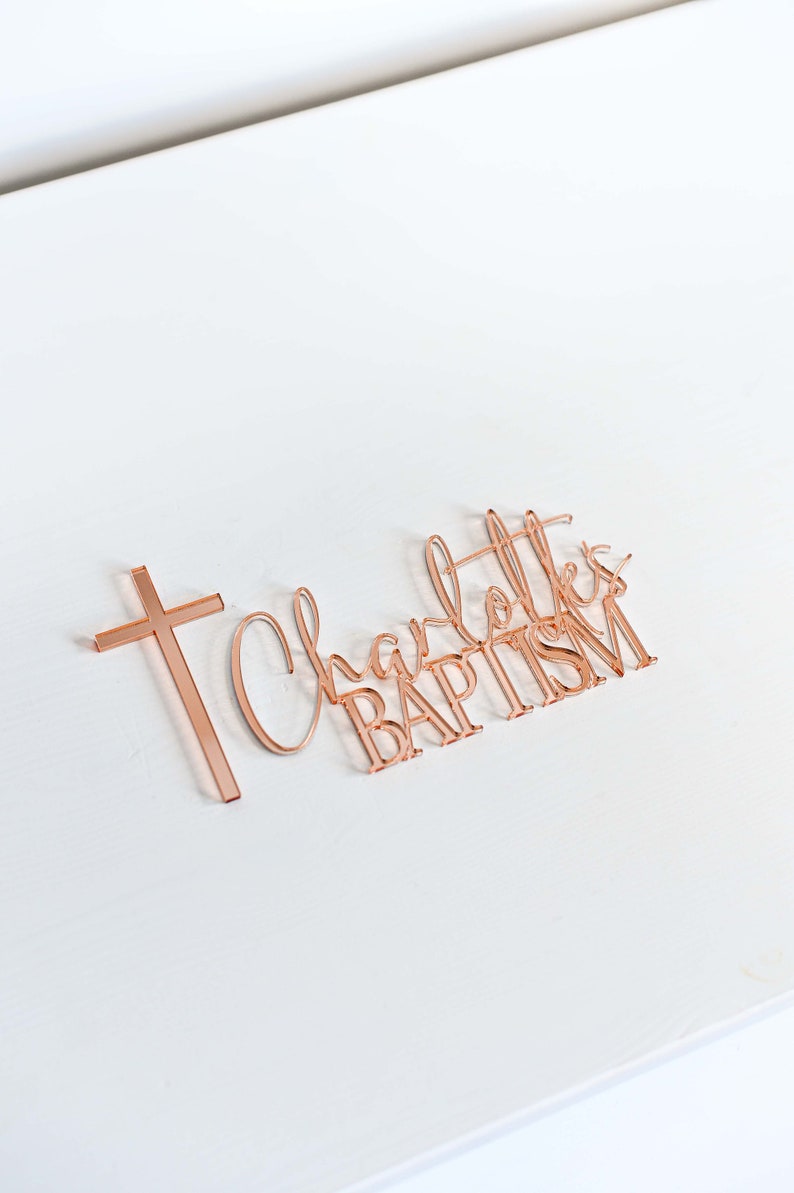 May include: Rose gold acrylic cake topper with the words "Charlotte's BAPTISM" in a script font, accompanied by a cross. The topper is placed on a white surface.