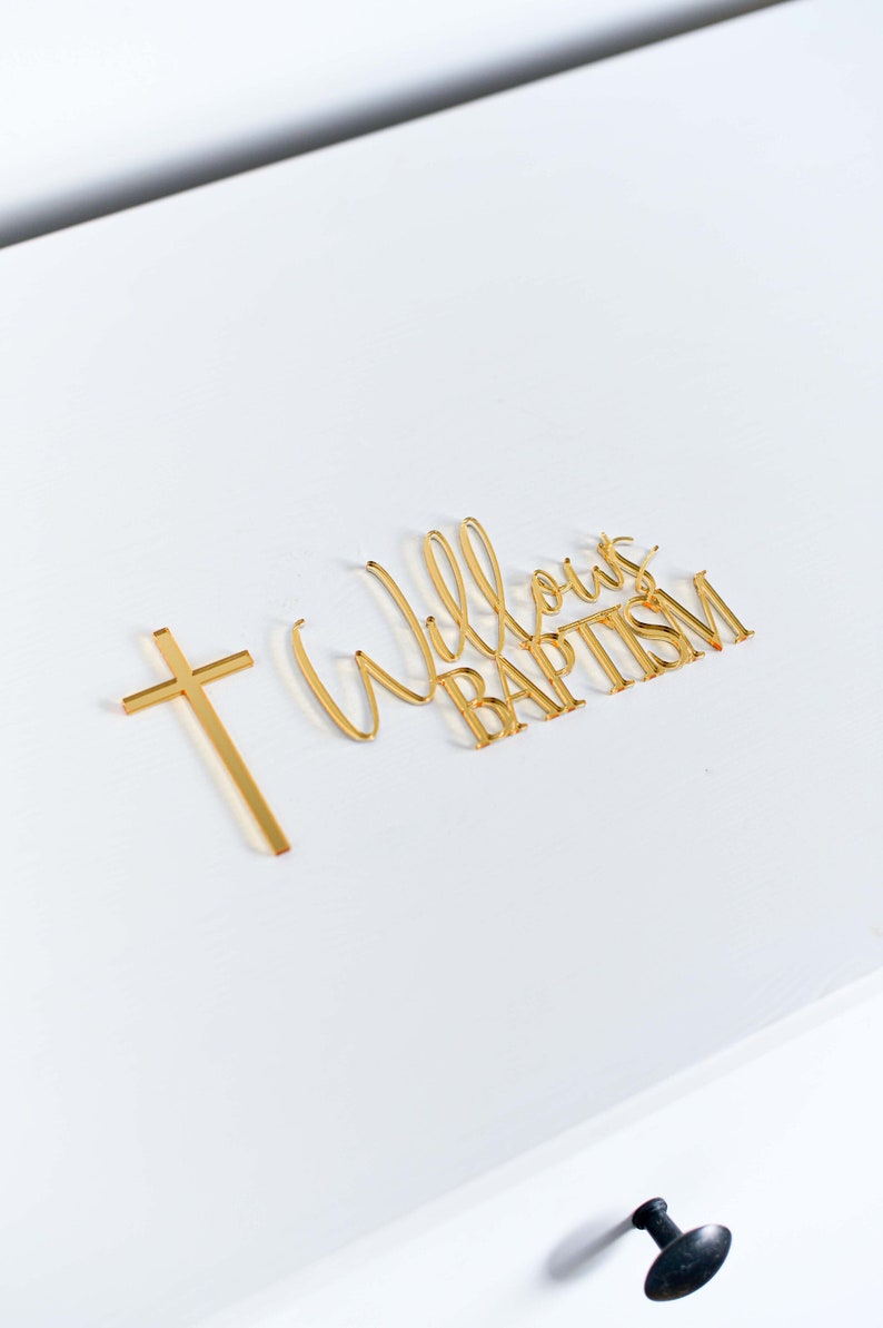 May include: Gold-colored baptism decorations on a white surface. The decorations include a cross and the words "Willow's Baptism" in a cursive font. The image is well-lit, highlighting the metallic sheen of the decorations.