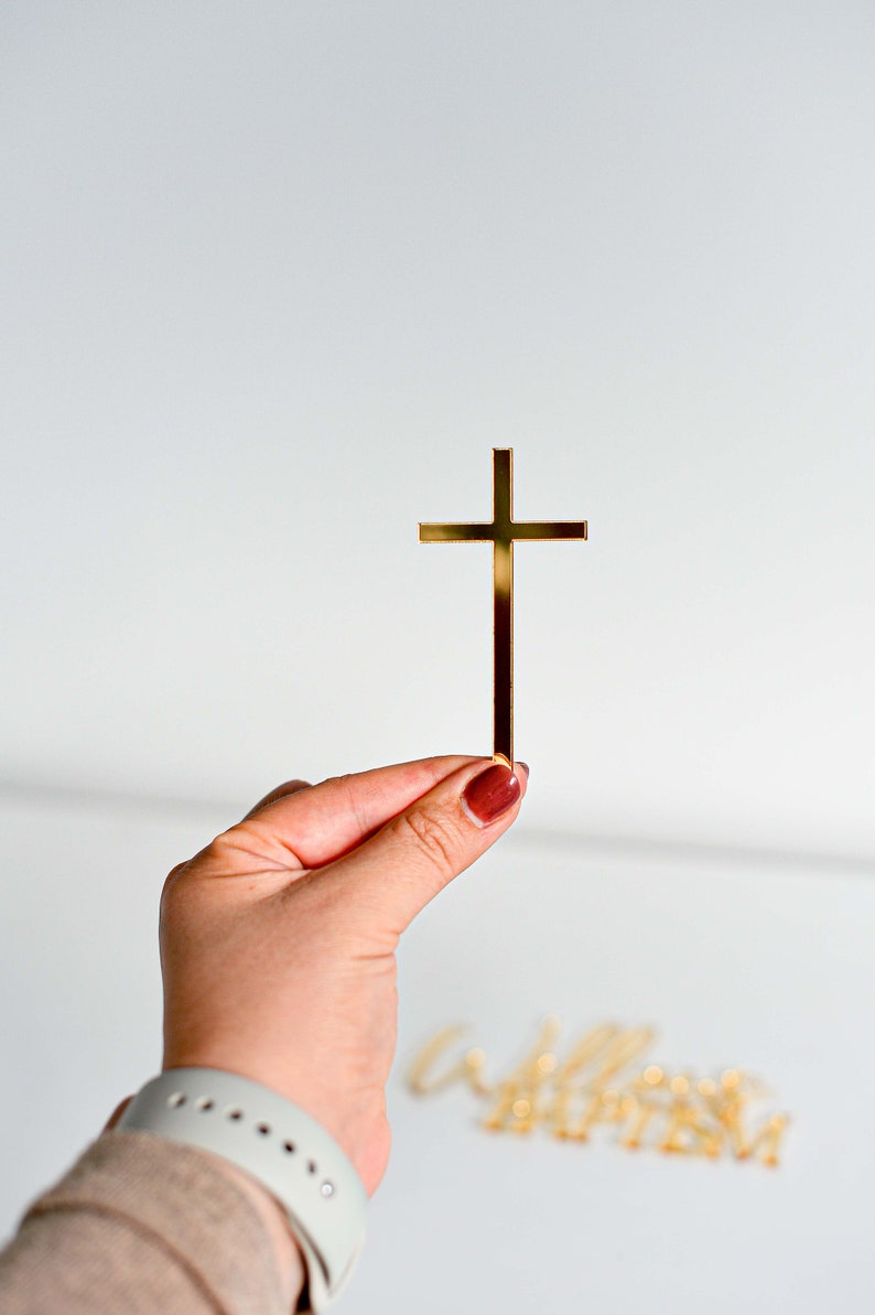 May include: A gold-colored cross held by a hand with a maroon nail polish. The cross is a simple, minimalist design. The background is a plain white surface. The word "welcome" is visible in the background.