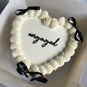 May include: A heart-shaped cake with white icing and piped buttercream edges. The word "engaged" is written in black script on the cake. Black satin ribbons are tied around the cake, which is in a white box.