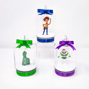 May include: Three clear glass jar ornaments with character figurines inside. Each jar has a colourful ribbon and bow. One features Woody, another Rex, and the third Buzz Lightyear. The ornaments are on a white surface.