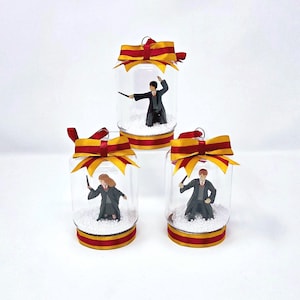 May include: Three clear glass jars with figurines inside, each adorned with a red and gold ribbon. The jars contain miniature figures of characters, set on a bed of white beads. The jars are decorated with red and gold ribbon.