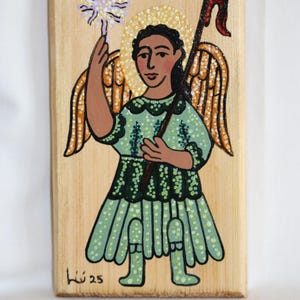 Puede incluir: Obra de arte pintada a mano sobre madera que representa un ángel con piel morena, vestido con un vestido verde y sosteniendo un bastón y una flor. El ángel tiene alas y un halo. La obra está firmada "Lú 25".