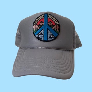 Peut inclure: Un chapeau de camionneur gris avec un patch en forme de signe de paix représentant une scène de montagne avec un lac bleu et un coucher de soleil rouge. Le patch est entouré d'un soleil jaune.