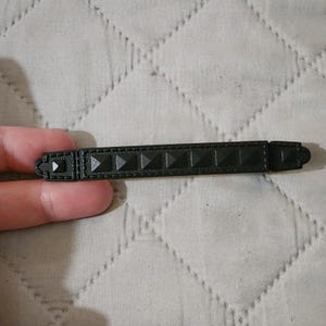 May include: A matte black hair barrette featuring a long, rectangular design with a series of raised pyramid-shaped studs running along the center. The accessory has a textured border and a secure clasp mechanism on the underside for hair styling.