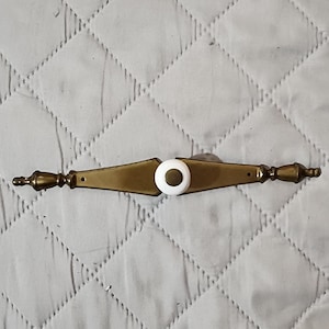 May include: A horizontal brass drawer pull featuring a central white ceramic knob with a brass center inlay. The metal hardware has a tapered, elongated diamond shape with decorative finials on each end and two visible screw holes for mounting.