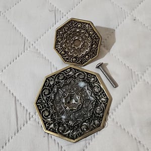 May include: Two octagonal brass-toned metal drawer pulls featuring intricate filigree patterns and a central faceted glass inlay. A single threaded metal mounting screw is included, positioned to the right of the larger decorative pull.