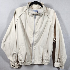 May include: A tan zip-up jacket with a stand-up collar, two front pockets, and elastic cuffs and hem. The jacket has a full-length zipper and a light-colored lining. The jacket is hanging on a black hanger.