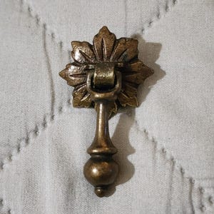 May include: A brass-toned metal drawer pull featuring a decorative backplate shaped like a multi-petaled flower. The hardware includes a hinged, vertical drop handle with a rounded, bulbous base, exhibiting a weathered, aged metallic finish.