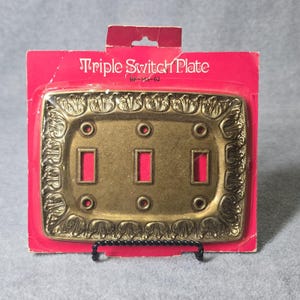 May include: A brass-toned metal triple switch plate with an ornate embossed border featuring leaf-like patterns. The rectangular plate has three vertical rectangular cutouts and six screw holes, packaged on a red cardboard backing labeled Triple Switch Plate BF-143-62.