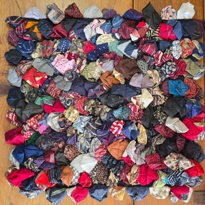 May include: A square textile wall hanging composed of numerous folded neckties in a variety of colors and patterns, including red, blue, black, white, and gold, arranged in a dense, overlapping grid pattern on a dark backing fabric.