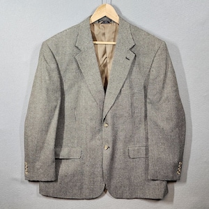 May include: A brown and white houndstooth blazer with a two-button closure, notched lapels, and three buttons on each cuff. The blazer is hanging on a wooden hanger against a gray background. The interior lining is a tan color.