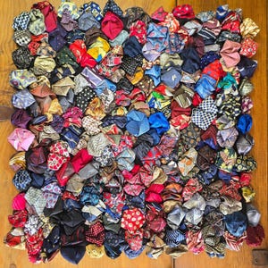 May include: A square textile wall hanging or mat composed of numerous folded fabric squares in a variety of colors and patterns, including polka dots, stripes, and geometric prints, arranged in a dense, multi-layered grid formation.