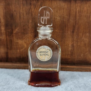 May include: A clear glass perfume bottle with a decorative stopper. The bottle has a label that reads "MOMENT SUPREME." The liquid inside is a dark amber color. The background is a wooden surface.