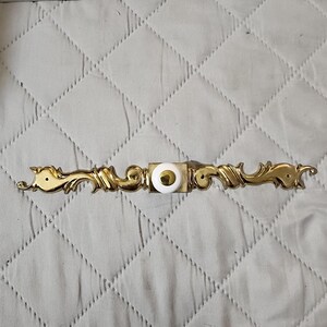 May include: A gold-toned metal drawer pull featuring an elongated, ornate scrollwork design with a central square base. The center of the pull is accented by a round, white ceramic knob with a small, circular gold-toned metal inlay at its peak.