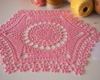 Puede incluir: Un tapete rosa hecho a mano, tejido a ganchillo con un diseño calado detallado. El tapete tiene un centro circular con un patrón de estrella y un borde festoneado. Bobinas de hilo y una pequeña ilustración de pájaro en el fondo.