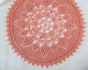 Puede incluir: Un tapete de ganchillo hecho a mano, de color coral, con un intrincado diseño floral en el centro. El tapete tiene un borde festoneado y se muestra sobre una superficie de madera clara. Esta pieza decorativa es ideal para añadir encanto vintage a cualquier hogar.
