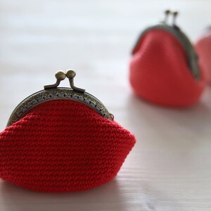 May include: Three crocheted coin purses with metal clasps. The purses are red, pink, and red. The purses are sitting on a white surface.