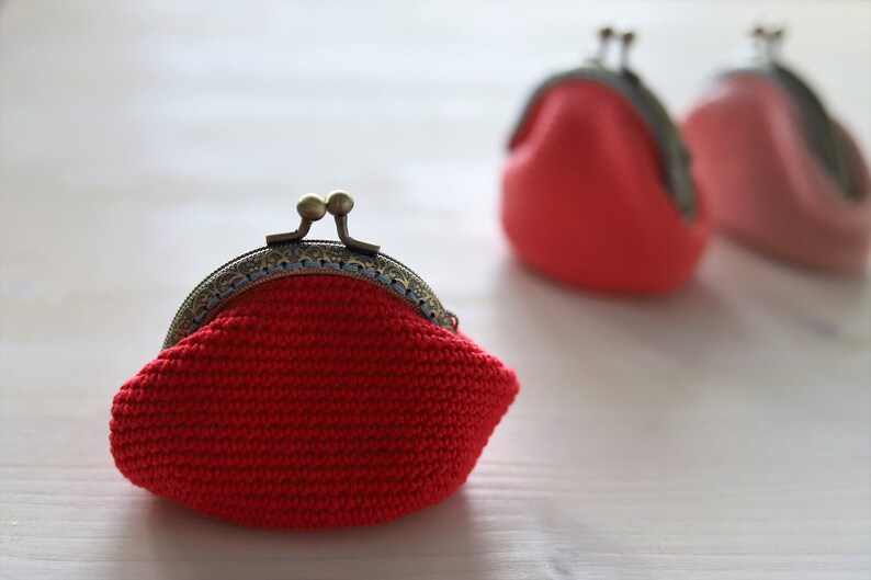 May include: Three crocheted coin purses with metal clasps. The purses are red, pink, and red. The purses are sitting on a white surface.